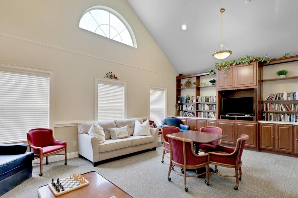 An indoor sitting area with white couch, a blue sofa chair, a brown table with pink cushioned chairs around it, all located in front of a long bookshelf full of books and a TV.