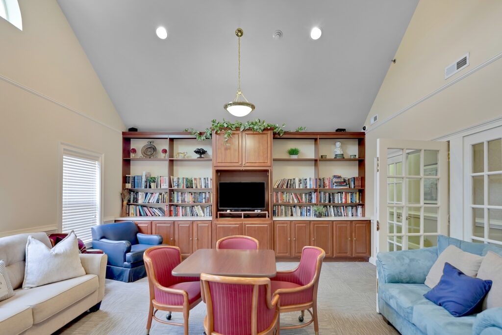 MG_7988 - The Willows of Easley An indoor sitting area with white and blue couches, a blue sofa chair, a brown table with pink cushioned chairs around it, all located in front of a long bookshelf full of books and a TV.