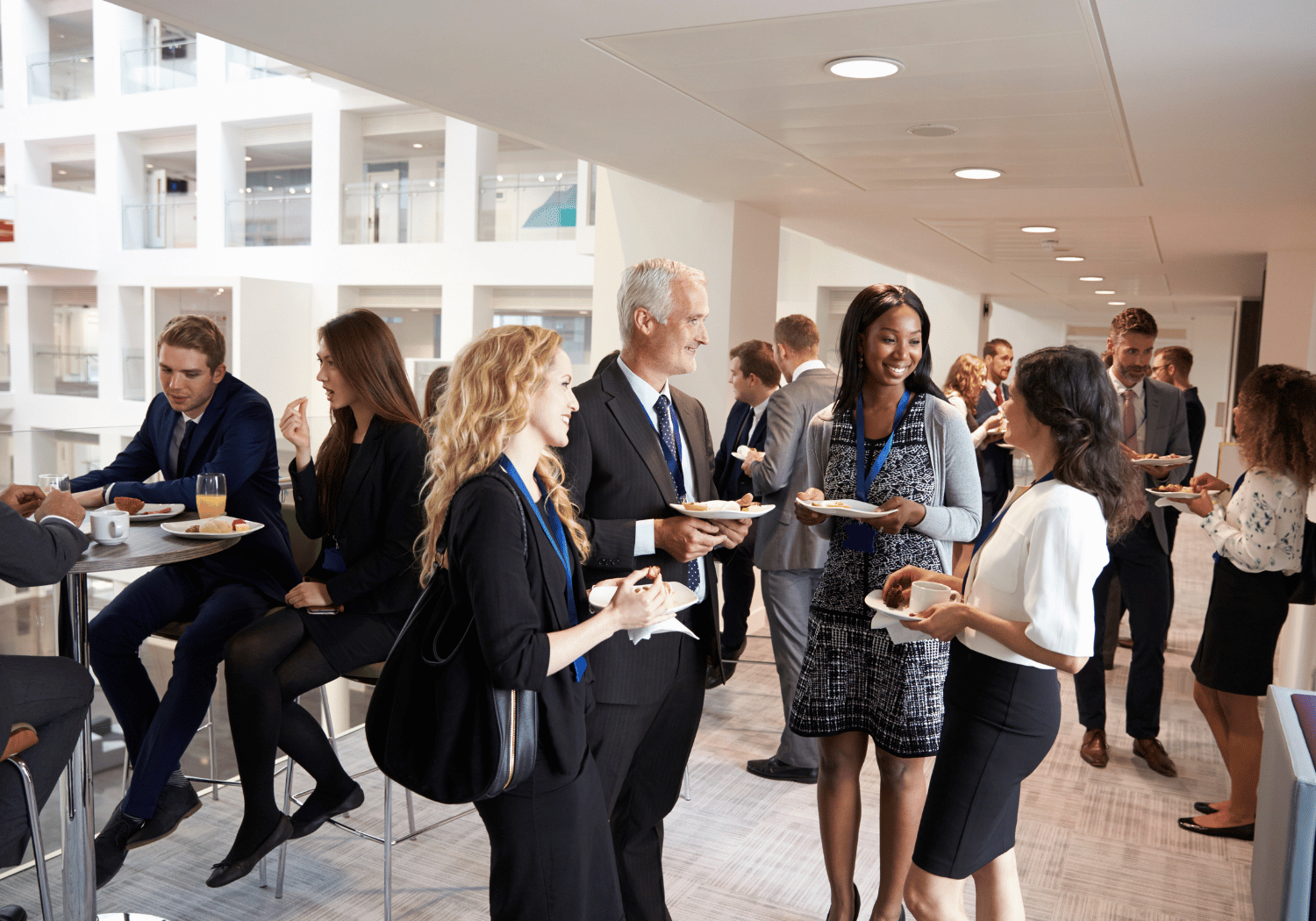 Professional networking event: people in business attire conversing and holding plates of food in a bright, modern office lobby.
